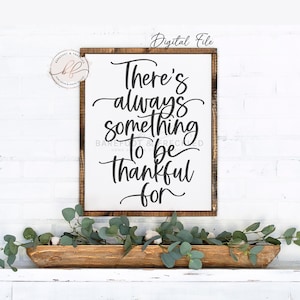 May include: A wooden framed sign with the words "There's always something to be thankful for" in black script. The sign is on a white brick wall, with a wooden trough filled with greenery below.