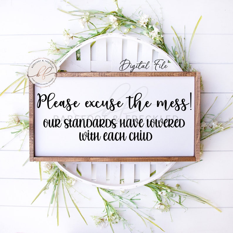 May include: A white wooden sign with brown trim, featuring the text "Please excuse the mess! OUR STANDARDS HAVE LOWERED with each CHILD". The sign is surrounded by white flowers and greenery.
