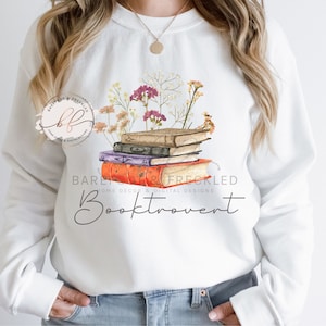 May include: White sweatshirt with a stack of books and dried flowers printed on the front. The text "Booktrovert" is printed below the books.