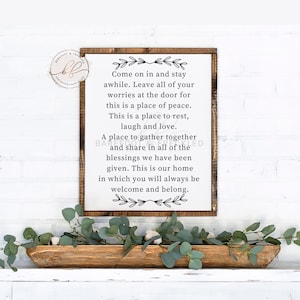 May include: A white sign with black text and a wood frame. The sign reads "Come on in and stay awhile. Leave all of your worries at the door for this is a place of peace. This is a place to rest, laugh and love. A place to gather together and share in all of the blessings we have been given. This is our home in which you will always be welcome and belong."