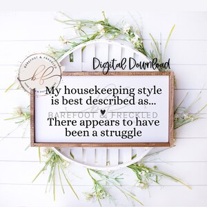 May include: A white wooden sign with the text "My housekeeping style is best described as... There appears to have been a struggle." The sign is framed in brown wood and has a heart symbol above the text "BAREFOOT & FRECKLED".
