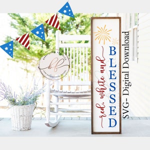 May include: A wooden sign with a red, white, and blue color scheme. The sign reads "red, white and BLESSED" with a firework graphic. The sign is framed in a dark wood frame. The sign is on a porch with a white rocking chair and a basket of flowers.