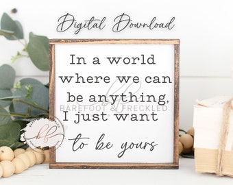 May include: A white sign with a wood frame reads "In a world where we can be anything, I just want to be yours". The sign is sitting on a table with a stack of books and a string of wooden beads.