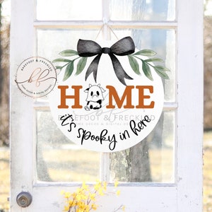 Può includere: Un appendiporta rotondo bianco con le parole "Home It's Spooky in here" scritte in nero e arancione. Un fiocco nero con foglie verdi è in cima all'appendiporta. Una piccola illustrazione di una mucca bianca e nera è al centro della parola "Home".