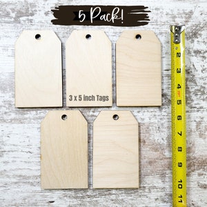 May include: Five unfinished wooden gift tags, each with a hole at the top. The tags are 3 inches by 5 inches and are arranged in a row on a white wooden surface. A yellow measuring tape is to the right of the tags.