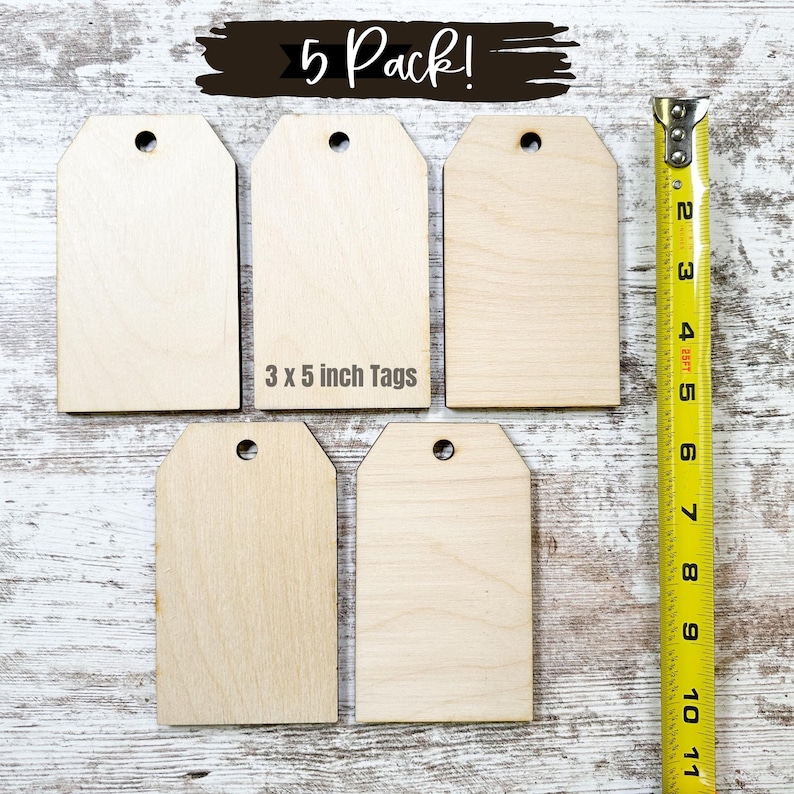 May include: Five unfinished wooden gift tags, each with a hole at the top. The tags are 3 inches by 5 inches and are arranged in a row on a white wooden surface. A yellow measuring tape is to the right of the tags.