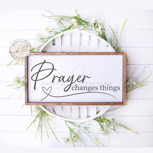 Peut inclure: Une pancarte en bois avec le texte "Prayer changes things" en lettres cursives brunes avec un petit cœur au-dessus du mot "Prayer". La pancarte est entourée de fleurs blanches et de verdure.