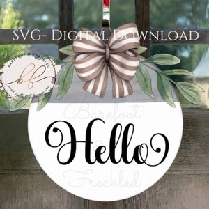 Op de afbeelding: Een witte ronde deurhanger met de tekst "Barefoot Hello Freckled" in zwarte cursieve letters. Een bruin-wit gestreepte strik met groene bladeren bevindt zich op de bovenste helft van de deurhanger.