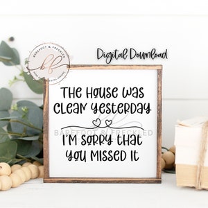 Könnte beinhalten: Ein weißes Schild mit braunem Holzrahmen, auf dem der Text "The house was clean yesterday. I'm sorry that you missed it." mit zwei Herzen über der zweiten Textzeile steht.