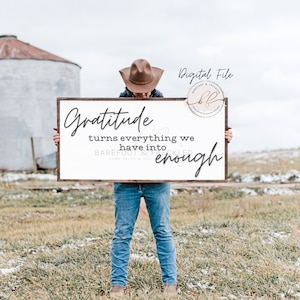 Könnte beinhalten: Ein weißes Schild mit braunem Holzrahmen, auf dem der Text "Gratitude turns everything we have into enough" steht. Das Schild wird von einer Person gehalten, die einen braunen Cowboyhut und eine blaue Jeans trägt.