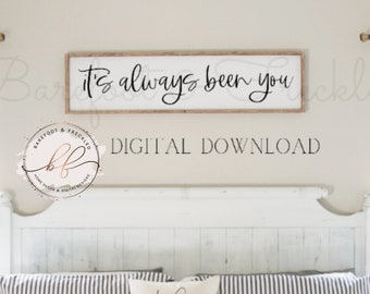 May include: A white wooden sign with black calligraphy that reads "It's always been you". The sign is hanging on a white wall above a white headboard with a striped bedspread.