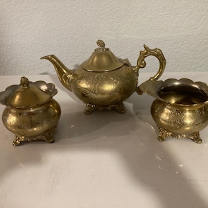 Könnte beinhalten: Ein goldfarbenes Teeset mit Teekanne, Milchkännchen und Zuckerdose. Jedes Stück ist mit einem verzierten Design versehen und steht auf drei Füßen.