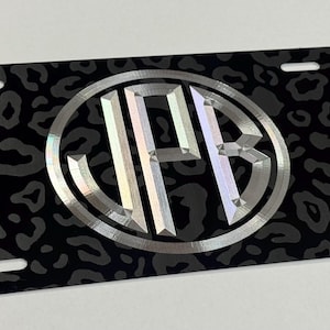 May include: Black license plate with a grey leopard print design. A silver monogram with the letters JPB is in the centre, within an oval. The plate has four mounting holes.