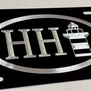 May include: Black license plate with a silver oval design. The design includes the letters "HH" and a lighthouse. The license plate has four mounting holes.