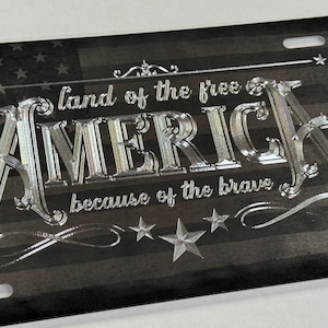 K&ouml;nnte beinhalten: Ein rechteckiges, schwarz-graues Nummernschild mit einem abgenutzten amerikanischen Flaggenhintergrund. Der Text "land of the free" und "because of the brave" umgibt das gro&szlig;e, metallisch gl&auml;nzende, gepr&auml;gte Wort "AMERICA" in der Mitte.