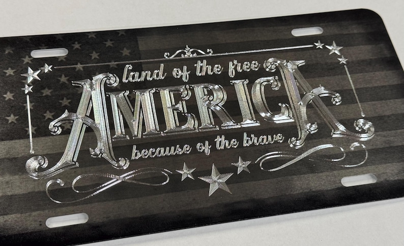 K&ouml;nnte beinhalten: Ein rechteckiges, schwarz-graues Nummernschild mit einem abgenutzten amerikanischen Flaggenhintergrund. Der Text "land of the free" und "because of the brave" umgibt das gro&szlig;e, metallisch gl&auml;nzende, gepr&auml;gte Wort "AMERICA" in der Mitte.