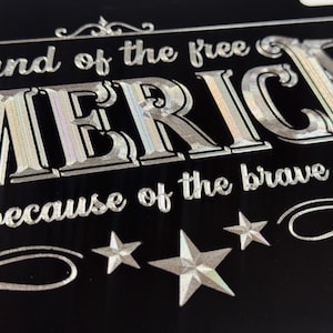 K&ouml;nnte beinhalten: Ein schwarzes rechteckiges Schild mit silbernem holografischem Text, der land of the free AMERICA because of the brave lautet. Das Design enth&auml;lt dekorative Schn&ouml;rkel und drei f&uuml;nfzackige Sterne am unteren Rand, alles in metallischer Optik.