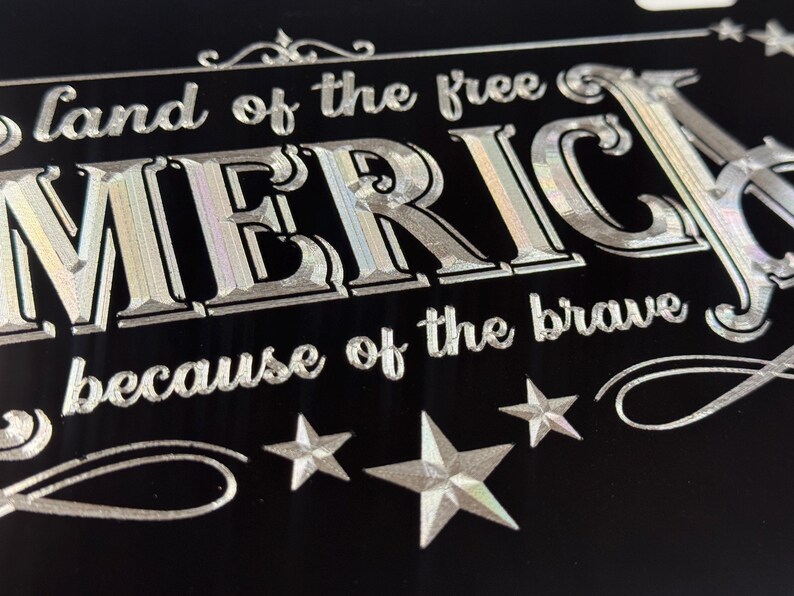 K&ouml;nnte beinhalten: Ein schwarzes rechteckiges Schild mit silbernem holografischem Text, der land of the free AMERICA because of the brave lautet. Das Design enth&auml;lt dekorative Schn&ouml;rkel und drei f&uuml;nfzackige Sterne am unteren Rand, alles in metallischer Optik.