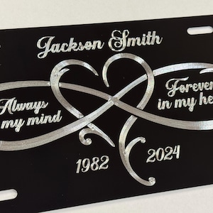May include: Black license plate with silver text and design. Features the name "Jackson Smith", the phrase "Always on my mind", and "Forever in my heart". Includes the years 1982 and 2024, and an infinity heart design.
