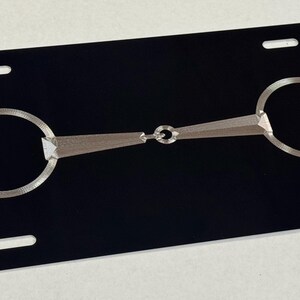 May include: A black license plate with a silver metal horse bit design. The bit features two circular rings connected by a central bar with a decorative element. The plate has four rectangular slots.