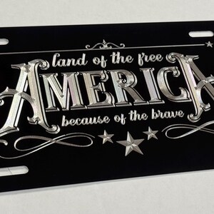 K&ouml;nnte beinhalten: Ein rechteckiges schwarzes Nummernschild mit silberner metallischer Schrift, die "land of the free" &uuml;ber dem Wort "AMERICA" und "because of the brave" darunter zeigt. Das Design enth&auml;lt dekorative silberne Sterne und Schn&ouml;rkel.