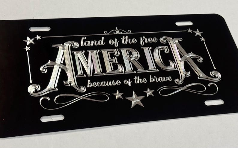 K&ouml;nnte beinhalten: Ein rechteckiges schwarzes Nummernschild mit silberner metallischer Schrift, die "land of the free" &uuml;ber dem Wort "AMERICA" und "because of the brave" darunter zeigt. Das Design enth&auml;lt dekorative silberne Sterne und Schn&ouml;rkel.