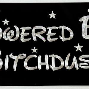 Könnte beinhalten: Schwarzes Nummernschild mit silberner Aufschrift "Powered By Bitchdust" und Sternen um den Text.