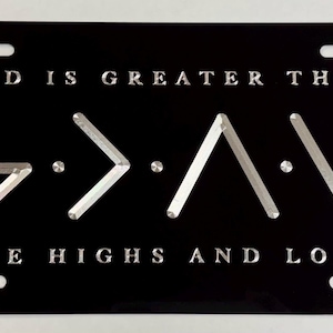 May include: Black license plate with the message "GOD IS GREATER THAN THE HIGHS AND LOWS" in silver lettering. The phrase "G > A > V" is also displayed in silver. The plate has four mounting holes.