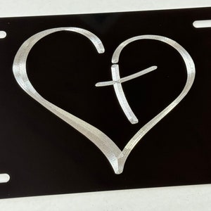 May include: A black metal license plate with a silver heart and a cross inside. The heart and cross are raised and have a textured finish.