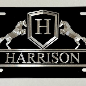 May include: Black license plate with a silver shield featuring the letter "H" and two silver horses on either side. The text "HARRISON" is below the shield.