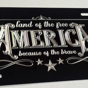 K&ouml;nnte beinhalten: Ein rechteckiges schwarzes Nummernschild mit silberner metallischer Schrift, die "land of the free" &uuml;ber dem Wort "AMERICA" und "because of the brave" darunter zeigt. Das Design enth&auml;lt dekorative silberne Sterne und Schn&ouml;rkel.