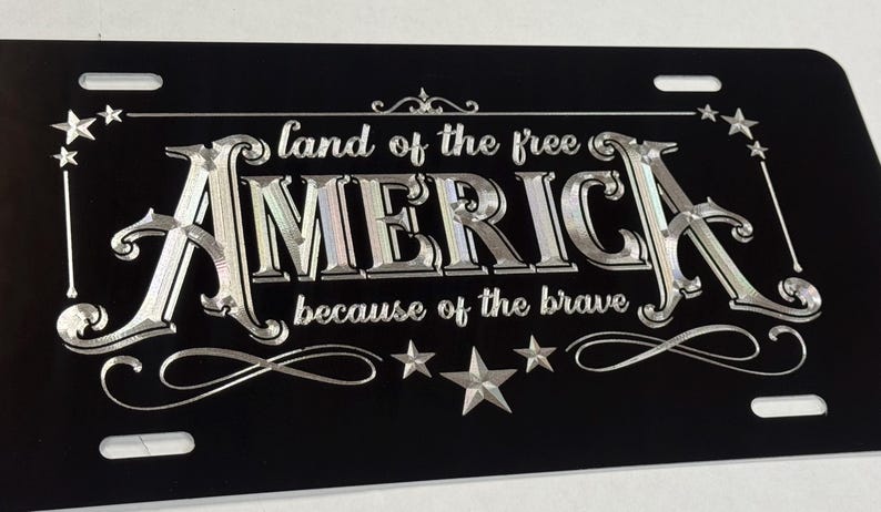 K&ouml;nnte beinhalten: Ein rechteckiges schwarzes Nummernschild mit silberner metallischer Schrift, die "land of the free" &uuml;ber dem Wort "AMERICA" und "because of the brave" darunter zeigt. Das Design enth&auml;lt dekorative silberne Sterne und Schn&ouml;rkel.