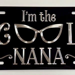 May include: Black license plate with the text "I'm the COOL NANA" in silver lettering. The word "COOL" is represented by a pair of eyeglasses. The license plate has four mounting holes.
