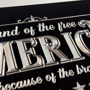 K&ouml;nnte beinhalten: Ein schwarzes, rechteckiges Nummernschild mit silberner, holografischer Schrift, die oben "land of the free", in der Mitte "AMERICA" in gro&szlig;en Gro&szlig;buchstaben und unten "because of the brave" zeigt, verziert mit kleinen Sternen.