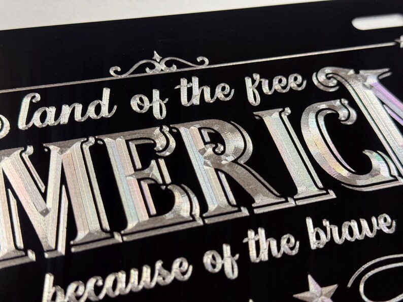 K&ouml;nnte beinhalten: Ein schwarzes, rechteckiges Nummernschild mit silberner, holografischer Schrift, die oben "land of the free", in der Mitte "AMERICA" in gro&szlig;en Gro&szlig;buchstaben und unten "because of the brave" zeigt, verziert mit kleinen Sternen.