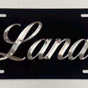 May include: Black license plate with the name "Lana" in a silver, cursive font. The plate has four mounting holes and a glossy finish. The name is the focal point, with a stylish, elegant design.