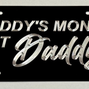 May include: Black license plate with the text "DADDY'S MONEY BUT I'M Daddy" in silver lettering. The plate has four mounting holes and a rectangular shape.