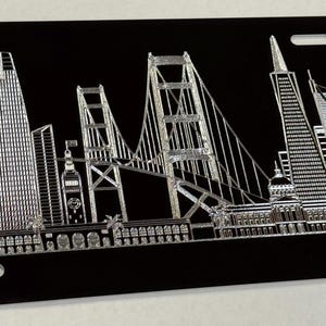 Puede incluir: Placa de matrícula negra con una representación plateada detallada del horizonte de San Francisco, incluyendo el Golden Gate Bridge y rascacielos. La placa tiene agujeros pre-perforados para el montaje.