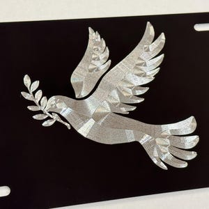 May include: A silver-colored metal dove with an olive branch in its beak is etched onto a black background. The dove is a symbol of peace.