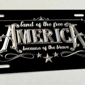 K&ouml;nnte beinhalten: Ein rechteckiges schwarzes Nummernschild mit silberner Metallschrift, die "land of the free AMERICA because of the brave" lautet. Das Design enth&auml;lt dekorative silberne Sterne und Schn&ouml;rkel sowie vier Befestigungsschlitze an den Ecken.