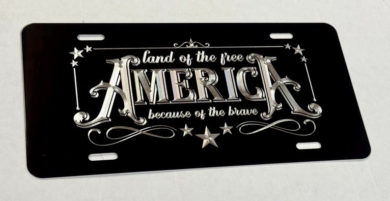 K&ouml;nnte beinhalten: Ein rechteckiges schwarzes Nummernschild mit silberner Metallschrift, die "land of the free AMERICA because of the brave" lautet. Das Design enth&auml;lt dekorative silberne Sterne und Schn&ouml;rkel sowie vier Befestigungsschlitze an den Ecken.