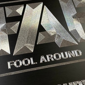 Pu&ograve; includere: Un cartello metallico argentato con la parola "FA" in grandi caratteri grassetto. Il testo "FOOL AROUND &" &egrave; stampato sotto le lettere.