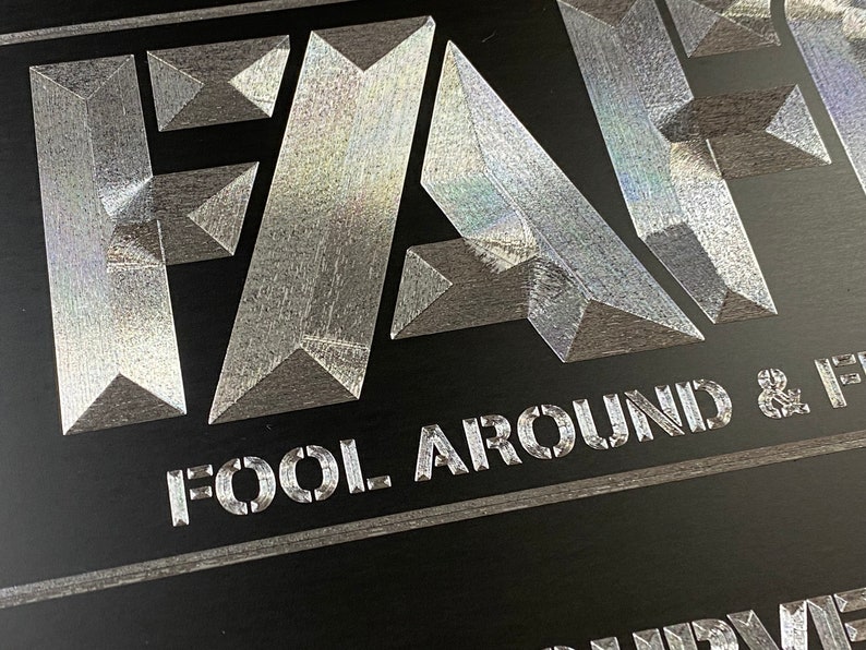 Pu&ograve; includere: Un cartello metallico argentato con la parola "FA" in grandi caratteri grassetto. Il testo "FOOL AROUND &" &egrave; stampato sotto le lettere.