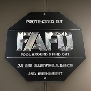 Pu&ograve; includere: Un cartello bianco e nero con il testo "Protected by FAFO Fool Around & Find Out 24 HR Surveillance 2nd Amendment" in un carattere metallico argentato.
