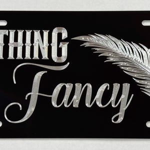 May include: Black metal license plate with silver lettering that reads "Nothing Fancy" and a silver feather design.