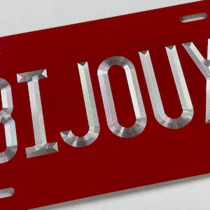 May include: A red license plate with the word "BIJOUX" in silver, embossed lettering. The plate has four rectangular slots, two at the top and two at the bottom. The word "BIJOUX" is a French word that means "jewels".