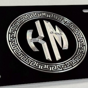 May include: Black license plate with a silver Greek key border and a central silver monogram. The monogram features the letters "KM" in a stylized, angular design. The plate has four mounting holes.
