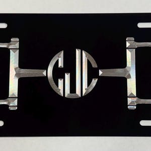 May include: Black license plate featuring a silver horse bit design with the initials GJC in the center. The plate has four mounting holes and a glossy finish, suitable for a vehicle.
