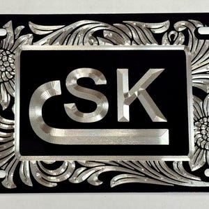 May include: A rectangular black license plate featuring a central silver-toned metallic inlay with the letters CSK. The plate is decorated with an intricate silver-toned floral and scrollwork pattern that covers the entire border area.