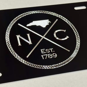 以下が含まれることがあります： 北カロライナ州のスタイリッシュなアウトライン、NCの文字、および「EST. 1789」のテキストを特徴とするシルバーサークルデザインのブラックナンバープレート。